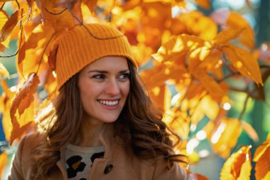 Hello autumn. happy modern 40 years old woman in beige coat and orange hat among autumnal foliage outside on the city park in autumn.