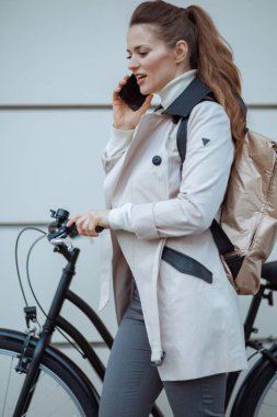 Bej renkli trençkotlu, bisiklet ve sırt çantalı modern bir kadın şehrin dışındaki metal bir duvarın önünde akıllı telefon kullanıyor..
