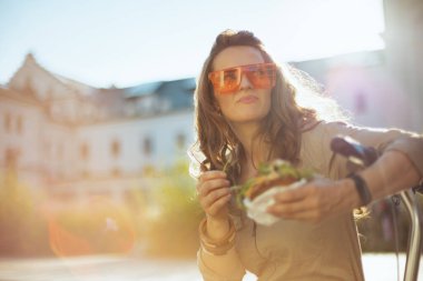 Gülümseyen 40 yaşında, güneş gözlüklü, mobilet ve sandviçli bir kadın şehirde oturuyor..