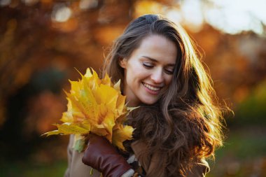 Merhaba Kasım ayı. Sonbaharda şehir parkında sonbahar sarısı yaprakları olan kahverengi ceketli, orta yaşlı, gülümseyen bir kadın..