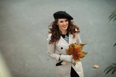 Merhaba Ekim. Sonbaharda şehrin dışında yeşil yaprakları olan bej renkli trençkot ve siyah bereli mutlu modern kadın manzarası..