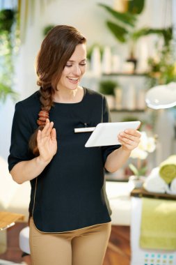 smiling middle aged woman worker with tablet PC talking video call in modern beauty studio.