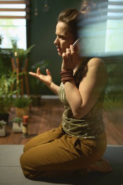 Modern yeşil oturma odasında, akıllı bir telefonla konuşan ve esneyen 40 yaşında modern bir kadın..