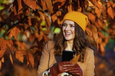 Hello november. happy stylish woman in brown coat and yellow hat using smartphone applications among autumnal foliage outside on the city park in autumn.