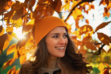 Hello september. happy modern middle aged woman in brown coat and yellow hat among autumnal foliage outdoors in the city park in autumn.
