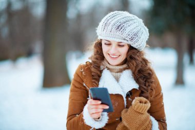 Örgü şapkalı ve koyun derisi ceketli gülümseyen modern kadın kışın şehir parkında akıllı telefon uygulaması kullanıyor..