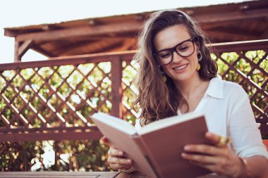 smiling modern woman in white shirt with eyeglasses sitting at the table reading book in the patio of guest house hotel.