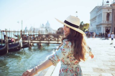 happy elegant tourist woman in floral dress with hat having walking tour on embankment in Venice, Italy.