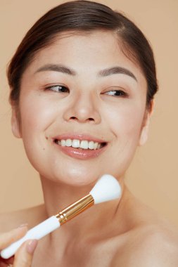 young asian woman with makeup brush against beige background.