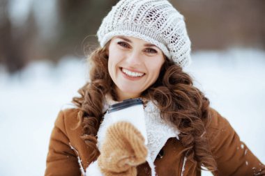 Kırk yıllık mutlu modern kadın portresi. Eldivenleri ve bir fincan sıcak çikolatası olan örgü şapkalı ve koyun derisi ceketli. Kışın şehir parkında..