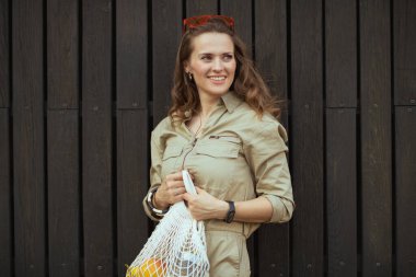 Orta yaşlı, modaya uygun bir kadın. İp torbası takmış, şehrin dışında siyah tahta duvara yaslanmış..