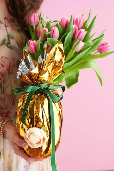 Closeup on woman with tulips bouquet and golden easter egg against pink background.