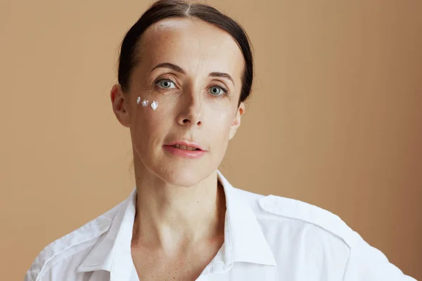 modern woman with eye cream on face in white shirt isolated on beige.