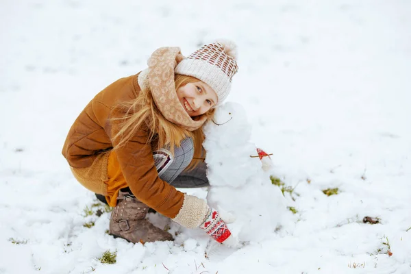 ニット帽とシープスキンのコートを着た幸せなスタイリッシュな女の子冬に市内公園でミトンと雪だるま屋外で — ストック写真