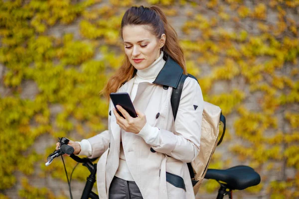 modern 40 years old woman in beige trench coat with bicycle and backpack using smartphone app against the green wall outside on the city street.