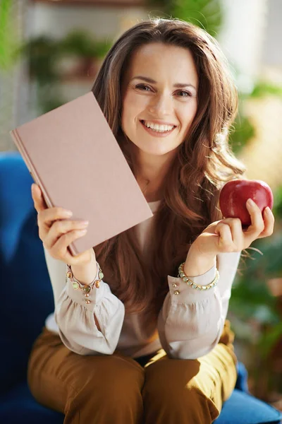 Green Home. smiling modern female with long wavy hair with book and red apple in the modern living room in sunny day.