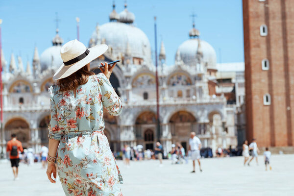 Seen from behind trendy traveller woman in floral dress with hat talking on a smartphone at Piazza San Marco in Venice, Italy.