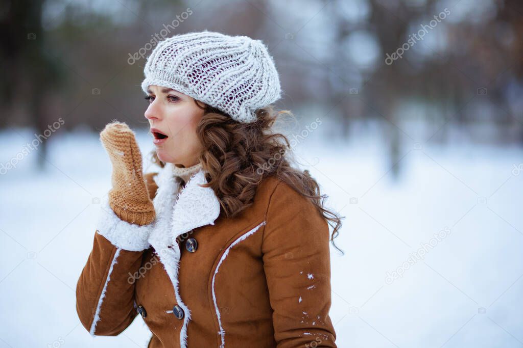 elegant female with mittens in a knitted hat and sheepskin coat coughing outdoors in the city park in winter.