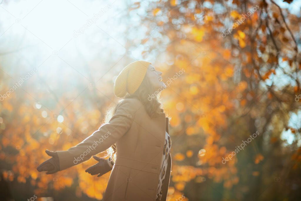 Hello october. smiling elegant female in brown coat and yellow hat rejoicing outside in the city park in autumn.