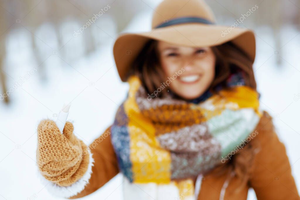 Closeup on smiling middle aged woman in brown hat and scarf with mittens and hygienic lipstick in sheepskin coat outside in the city park in winter.