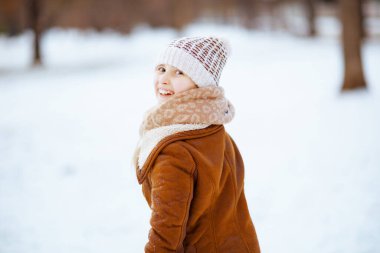 Güler yüzlü modern çocuk kışın şehir parkında örgü bir şapka ve koyun derisi ceketiyle oyun oynuyor..