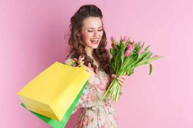 smiling trendy woman in floral dress with tulips bouquet and shopping bags against pink background.