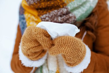 Closeup on middle aged woman in brown hat and scarf with mittens and napkin in sheepskin coat outdoors in the city park in winter.