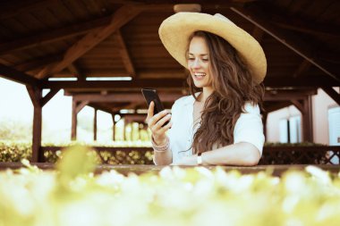 happy elegant woman in white shirt with hat sending text message using smartphone on the ranch.