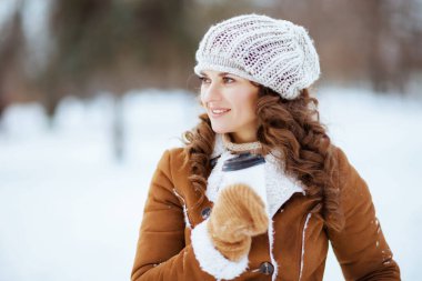 Mutlu, orta yaşlı bir kadın, eldivenleri ve bir fincan kapuçinosu olan, örgü şapkalı ve koyun derisi ceketli, kışın şehir parkında..