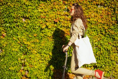 Güneş gözlüklü, spor çantalı ve scooterlı zarif bir kadın şehrin dışında yeşil duvara yaslanıyor..