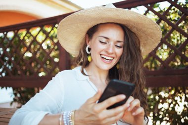 Beyaz tişörtlü, şapkalı mutlu modern kadın verandadaki akıllı telefonda görüntülü sohbet ediyor..