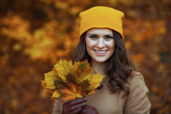 Hello september. smiling stylish woman in brown coat and yellow hat with autumn yellow leaves outdoors in the city park in autumn.