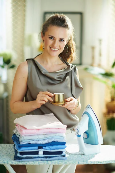 Happy modern woman in silk blouse and beige pants with steam iron, ironing board and pile of folded ironed clothes drinking cup of espresso after housework in the modern house in sunny day.