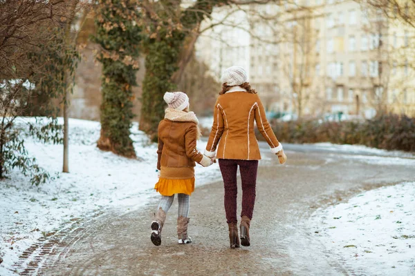 ニット帽と羊皮のコートを着た母親と子供の後ろから見ると 冬にはニット帽と羊皮のコートが市内公園の屋外を歩く — ストック写真