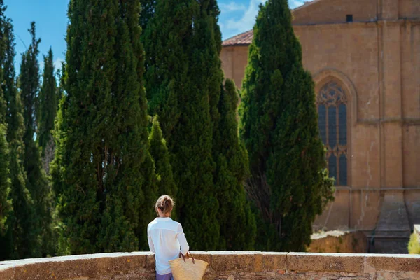 Travel in Italy. Seen from behind middle aged traveller woman with straw bag sightseeing near cathedral in Pienza in Tuscany, Italy.