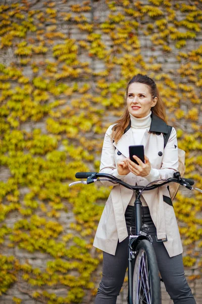 pensive modern woman in beige trench coat with bicycle using smartphone applications against the green wall outside in the city.