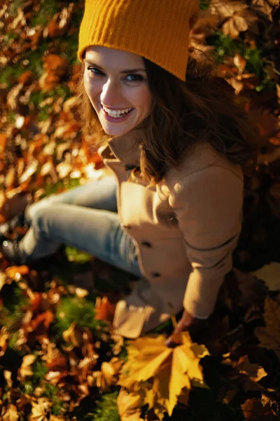 Hello november. Upper view of smiling young woman in brown coat and yellow hat with autumn yellow leaves sitting outdoors in the city park in autumn.