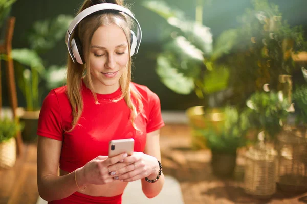 Uśmiechnięta Młoda Kobieta Czerwonym Stroju Fitness Smartfonem Słuchawkami Nowoczesnym Zielonym — Zdjęcie stockowe