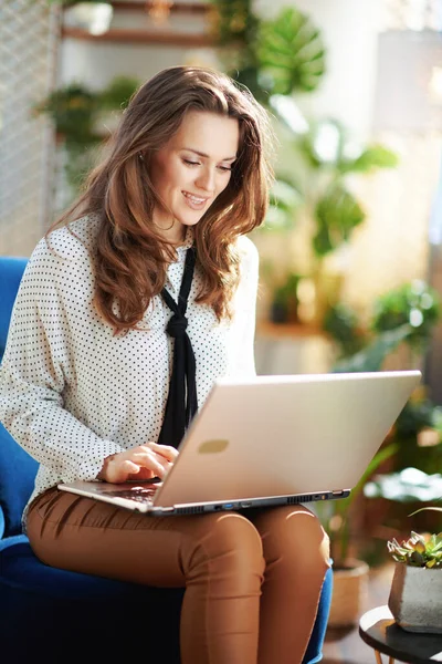 Green Home. happy trendy middle aged housewife with long wavy hair with laptop in beige pants and blouse in the modern living room in sunny day.