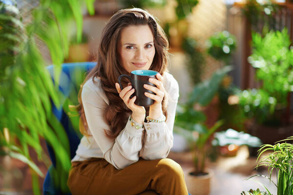 Green Home. Portrait of smiling young housewife with long wavy hair with cup of coffee in green pants and grey blouse in the modern house in sunny day.
