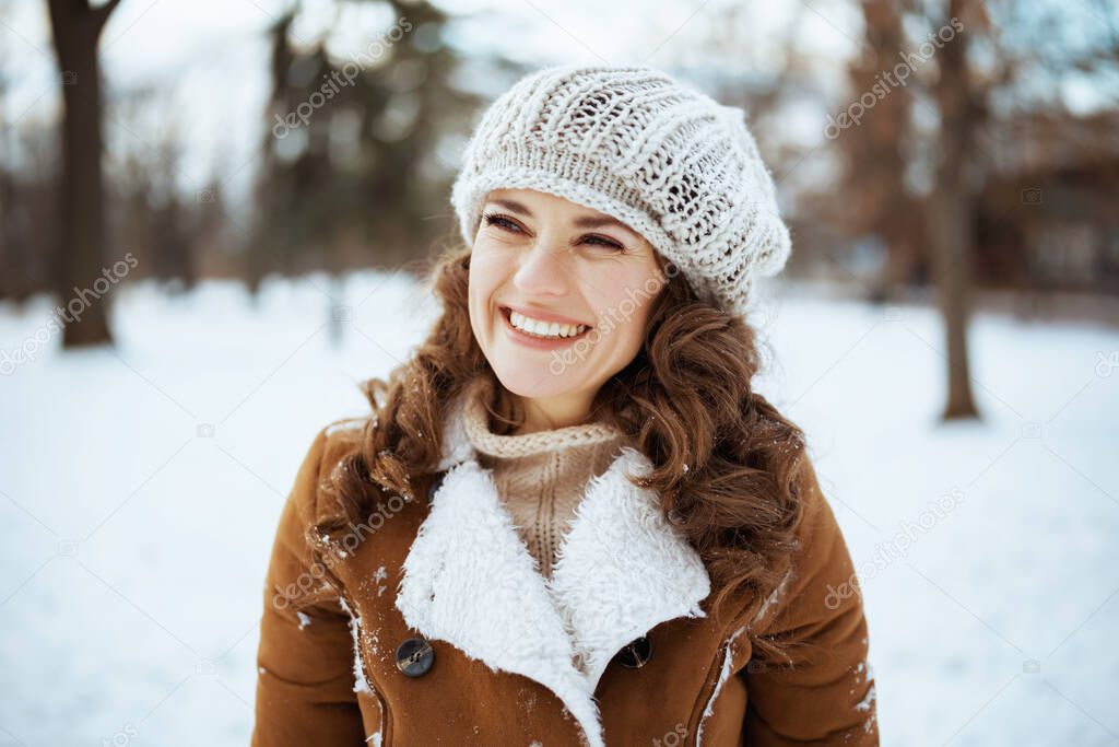 smiling modern 40 years old woman outdoors in the city park in winter in a knitted hat and sheepskin coat looking into the distance.