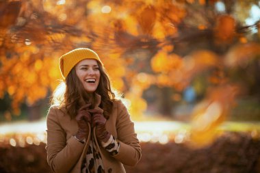 Hello autumn. happy stylish female in beige coat and orange hat outside in the city park in autumn.