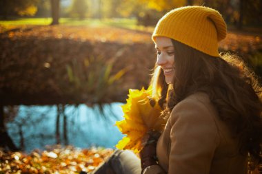 Merhaba Ekim. Sonbahar sarı yapraklı bej ceketli ve turuncu şapkalı gülümseyen modern kadın sonbaharda şehir parkında oturuyor..