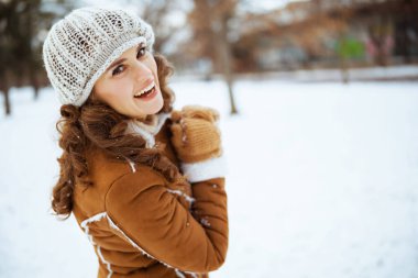 smiling elegant middle aged woman with mittens in a knitted hat and sheepskin coat outside in the city park in winter.