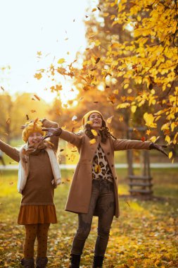 Merhaba Eylül. Sarı şapkalı modern anne ve kız güler yüzlü sonbaharda şehir parkında sonbahar yapraklarını fırlatır..