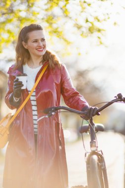happy modern middle aged woman in red rain coat with bicycle and cup of tea walking outside in the city.