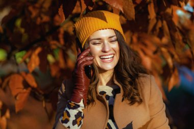 Hello november. smiling modern middle aged woman in beige coat and orange hat talking on a smartphone among autumnal foliage outside on the city park in autumn.