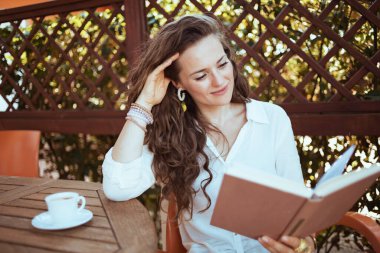 relaxed elegant female in white shirt with cup of coffee sitting at the table reading book in the terrace of guest house hotel.