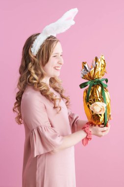 Uzun dalgalı sarı saçlı, tavşan kulaklı mutlu modern çocuk ve pembe arka planda altın paskalya yumurtası..