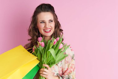 smiling trendy female in floral dress with tulips bouquet and shopping bags isolated on pink background.